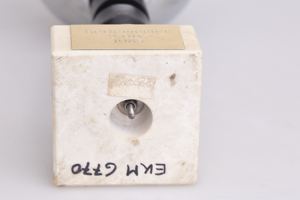 Pokal med halvrund topp, tre svarte runde ringer rundt stetten og en fot i hvit sten. Innskriften er "Seriemester 2. div. 1985"