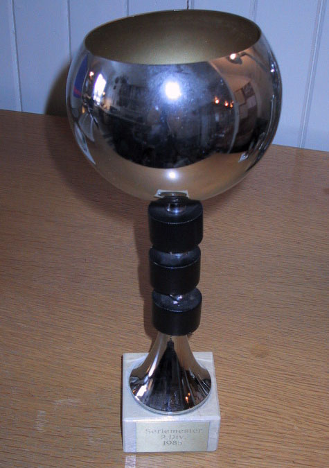 Pokal med halvrund topp, tre svarte runde ringer rundt stetten og en fot i hvit sten. Innskriften er "Seriemester 2. div. 1985"