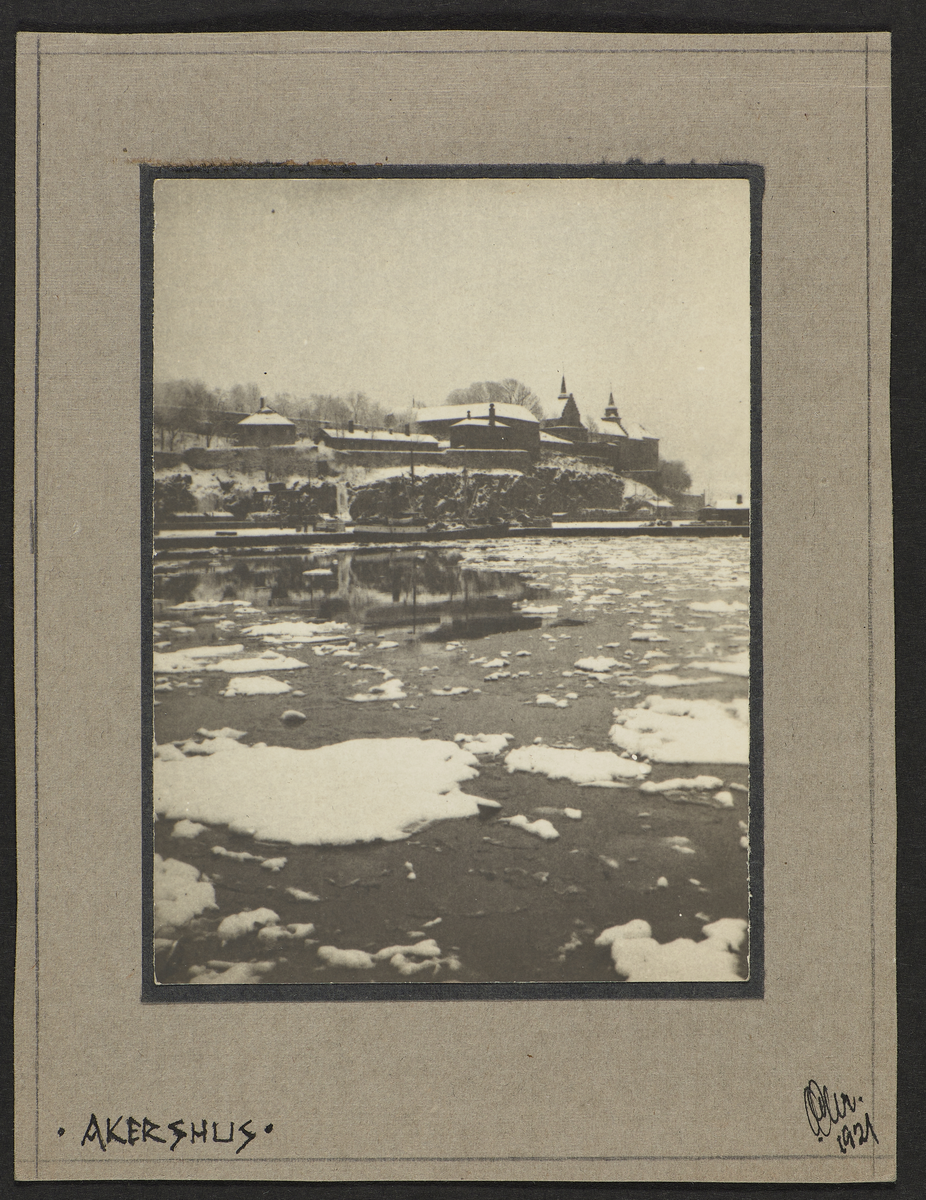Et vinterlandskap med utsikt over en isbelagt fjord til Akershus festning på en høyde. Et lag snø dekker alle tak og flater, og bak bygningen står det flere trær.
