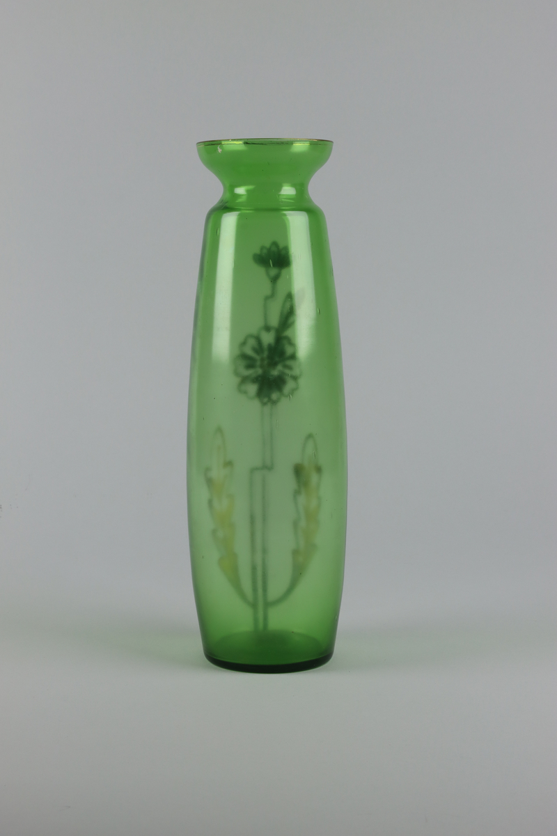 Vas av grönt glas. Spolformad kropp med en utvikt mynning upptill. Mynningen dekorerad med en förgylld kant och smal bård. Mitt fram på vasen dekor med blommor, stjälkar och blad i förgylld emalj. Allt dekor i guld och vitt mot en grön grund.