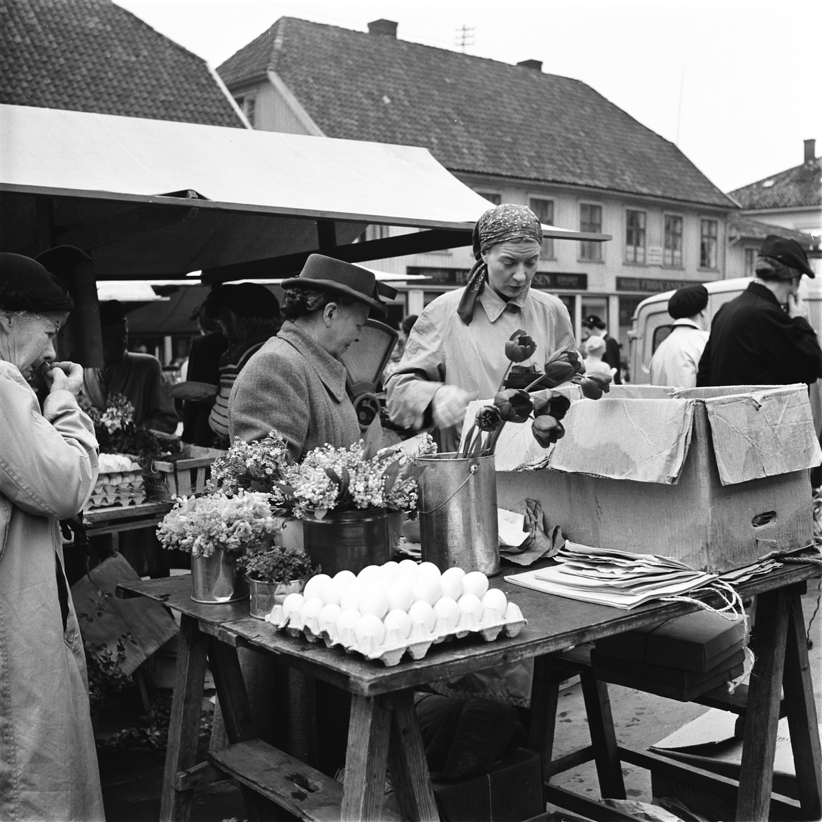 Livlig handel på torget i Tønsberg. I bildet ser vi blomsterpotter, kartonger med egg og en eske, og i bakgrunnen er det noen butikker.