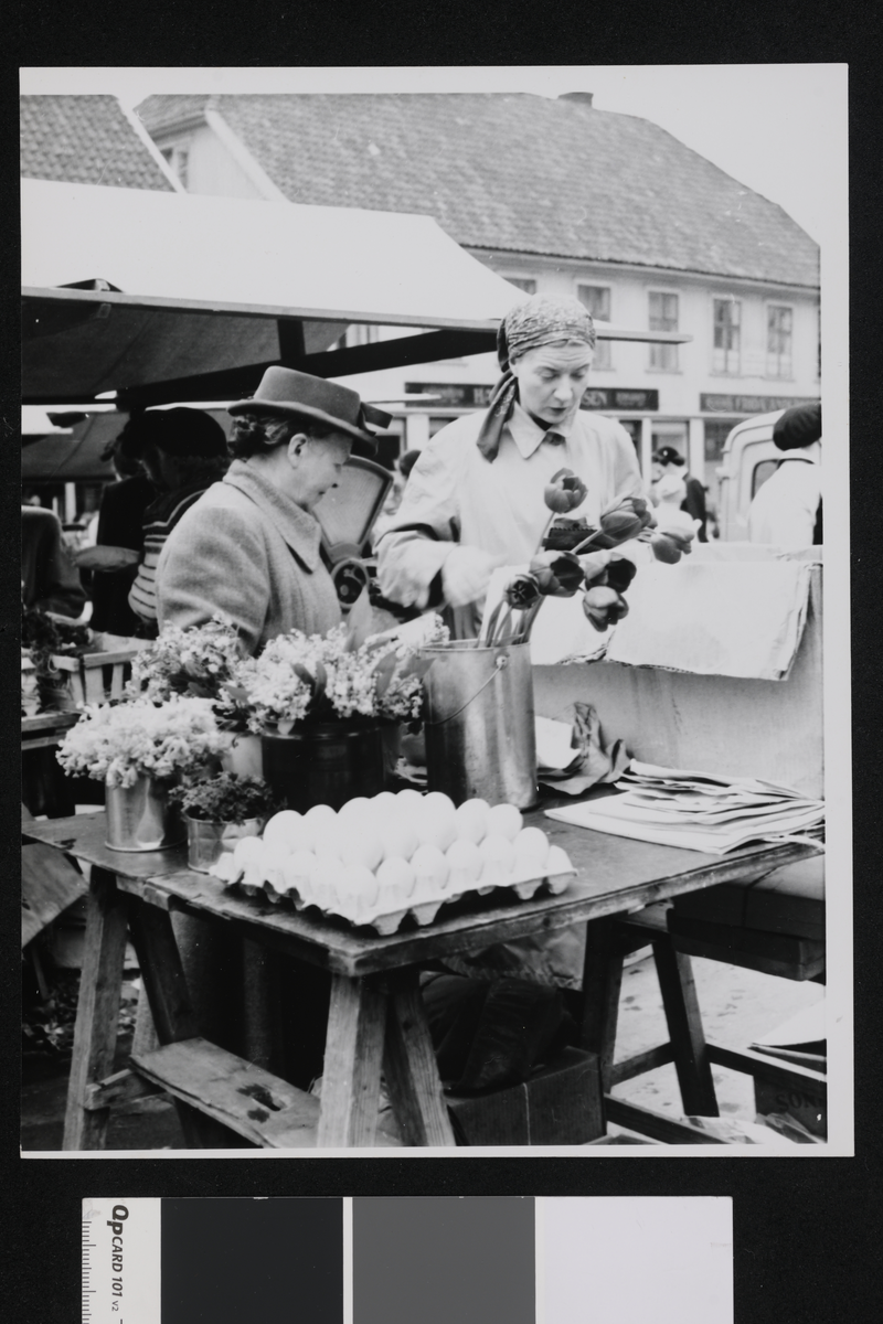 Livlig handel på torget i Tønsberg. I bildet ser vi blomsterpotter, kartonger med egg og en eske, og i bakgrunnen er det noen butikker.