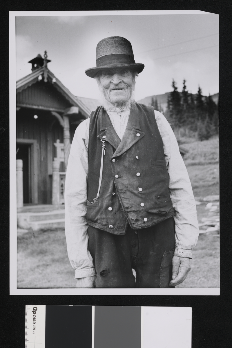 En eldre mann med hatt, vest og hvit skjorte. I bakgrunnen er det en trebygning som er en del av en gård, og en rekke med grantrær.
