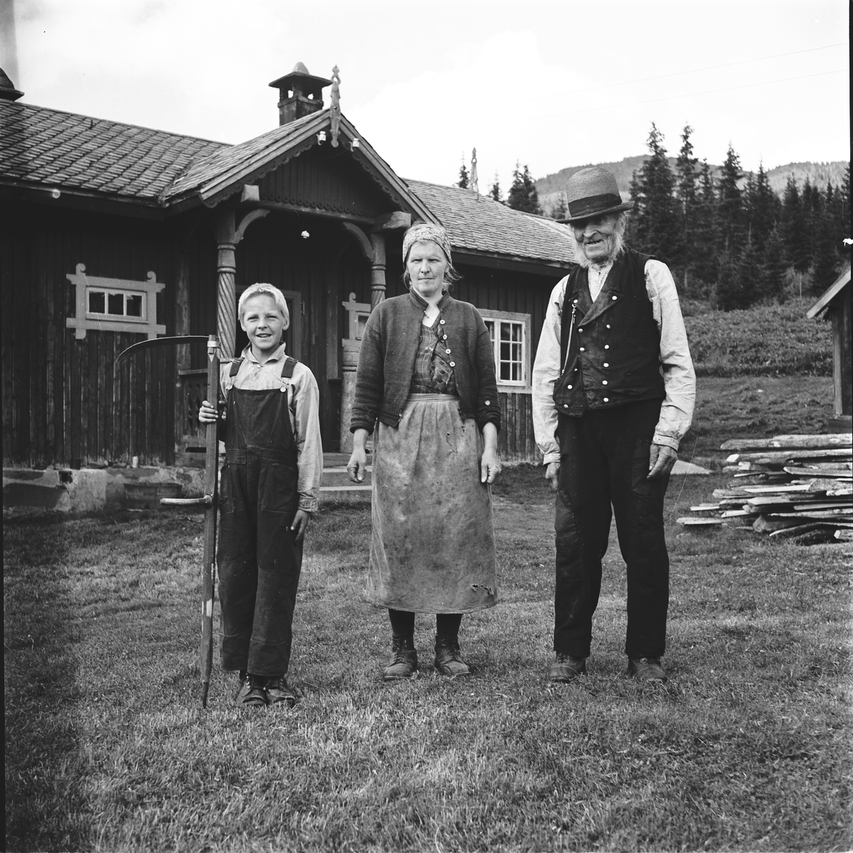 En familie i tre generasjoner på en gård. I bakgrunnen er det en mørk trebygning med søyler ved inngangspartiet, grantrær og fjell. På bildet ser vi Ola Olsen Guroplass, 96 år, svigerdatter og sønnesønn Ola H. Solbø 9 år. De tre drev denne gården da sønnen til den gamle var død.