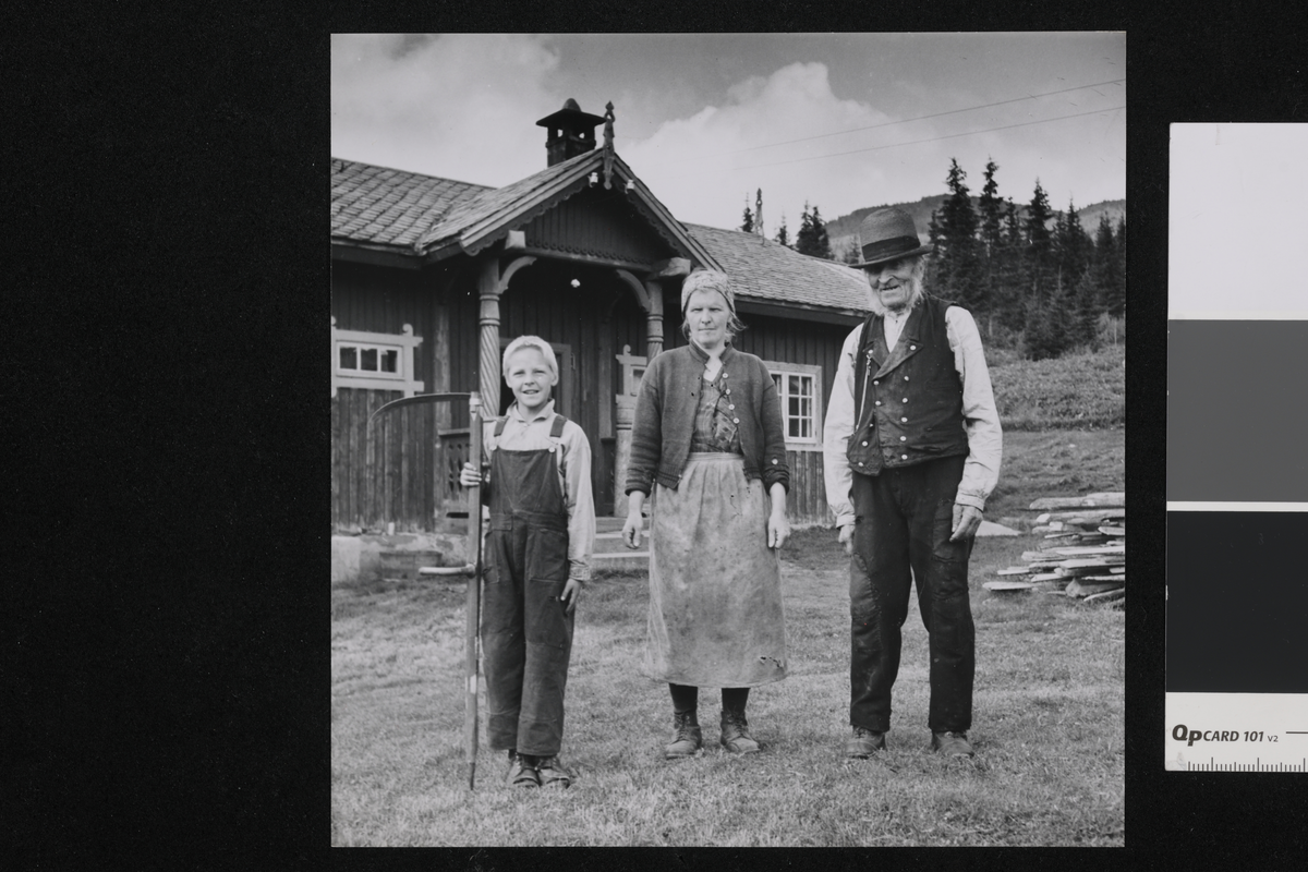 En familie i tre generasjoner på en gård. I bakgrunnen er det en mørk trebygning med søyler ved inngangspartiet, grantrær og fjell. På bildet ser vi Ola Olsen Guroplass, 96 år, svigerdatter og sønnesønn Ola H. Solbø 9 år. De tre drev denne gården da sønnen til den gamle var død.