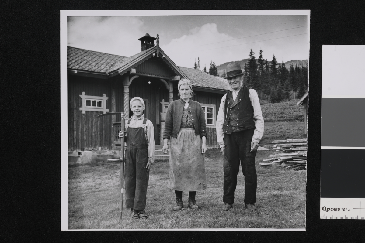 En familie i tre generasjoner på en gård. I bakgrunnen er det en mørk trebygning med søyler ved inngangspartiet, grantrær og fjell. På bildet ser vi Ola Olsen Guroplass, 96 år, svigerdatter og sønnesønn Ola H. Solbø 9 år. De tre drev denne gården da sønnen til den gamle var død.