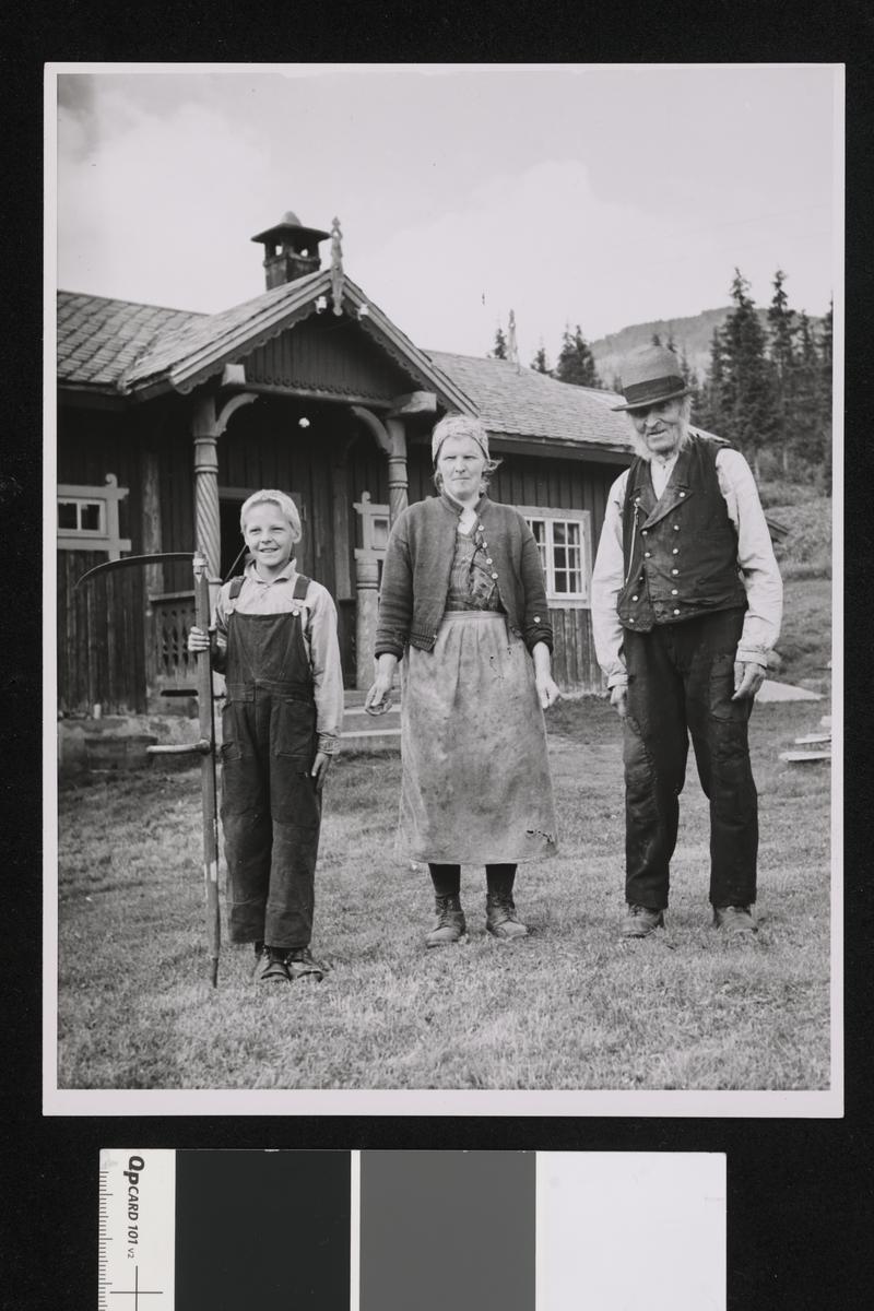 En familie i tre generasjoner på en gård. I bakgrunnen er det en mørk trebygning med søyler ved inngangspartiet, grantrær og fjell. På bildet ser vi Ola Olsen Guroplass, 96 år, svigerdatter og sønnesønn Ola H. Solbø 9 år. De tre drev denne gården da sønnen til den gamle var død.