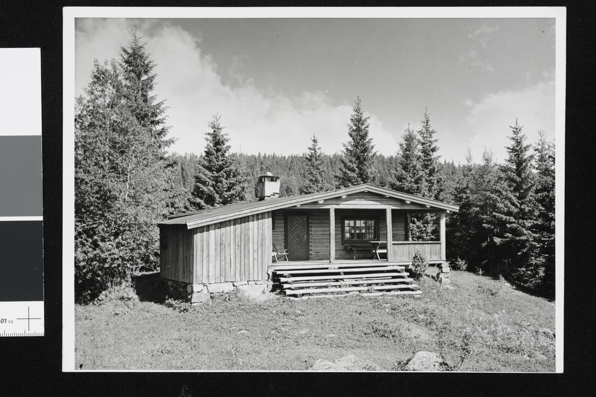 En trehytte på en høyde i en tett granskog. Hytten har terrasse og skrånende tak med pipe.