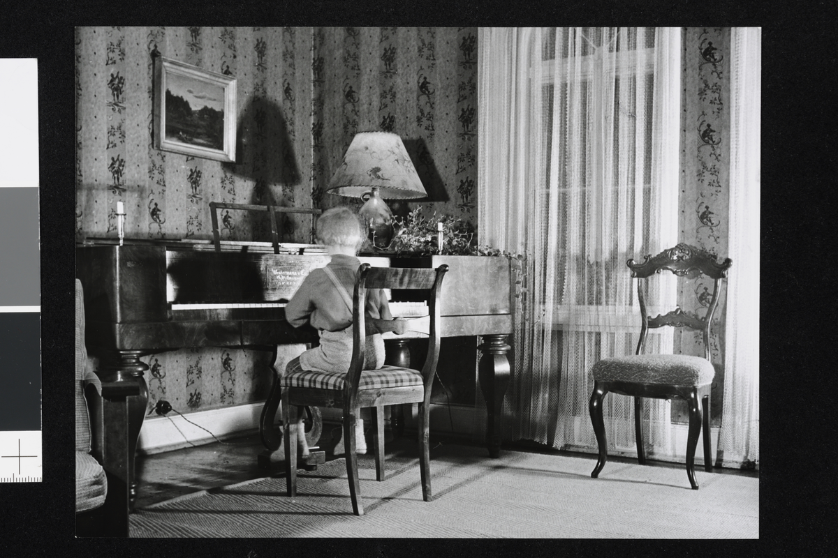 Et barn sitter ryggvendt ved et piano. Rommet er dekorert med mønstret tapet, en lett gardin, en bordlampe med skjerm og elegant utformede stoler.