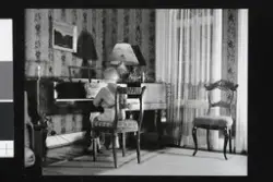 Et barn sitter ryggvendt ved et piano. Rommet er dekorert med mønstret tapet, en lett gardin, en bordlampe med skjerm og elegant utformede stoler.