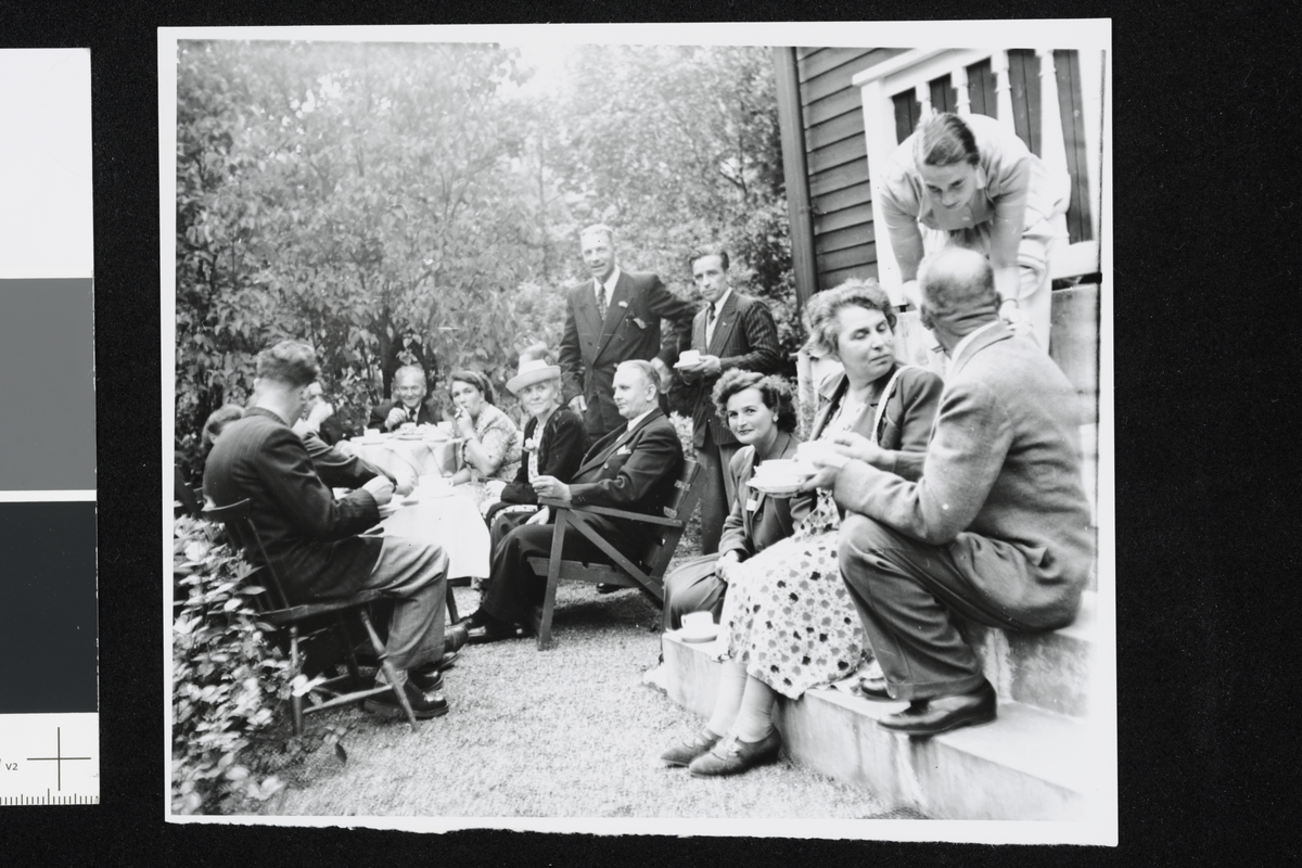 En gruppe med mennesker drikker kaffe i hagen. Antatt fra en serie bilder Meyer tok av flyktninger (polske, tsjekkiske og jugoslaviske) som er på besøk hos Alfhild Myhre. Se liten sak i Urd nr. 18. 54 årg. 26. august 1950.