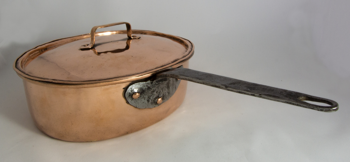 Kastrull med lock tillverkad i koppar. Oval form med raka sidor. Lockets handtag har hjärtbladsformade fästen. Kastrullen har ett handtag av järn med ett halvcirkelformat fäste.