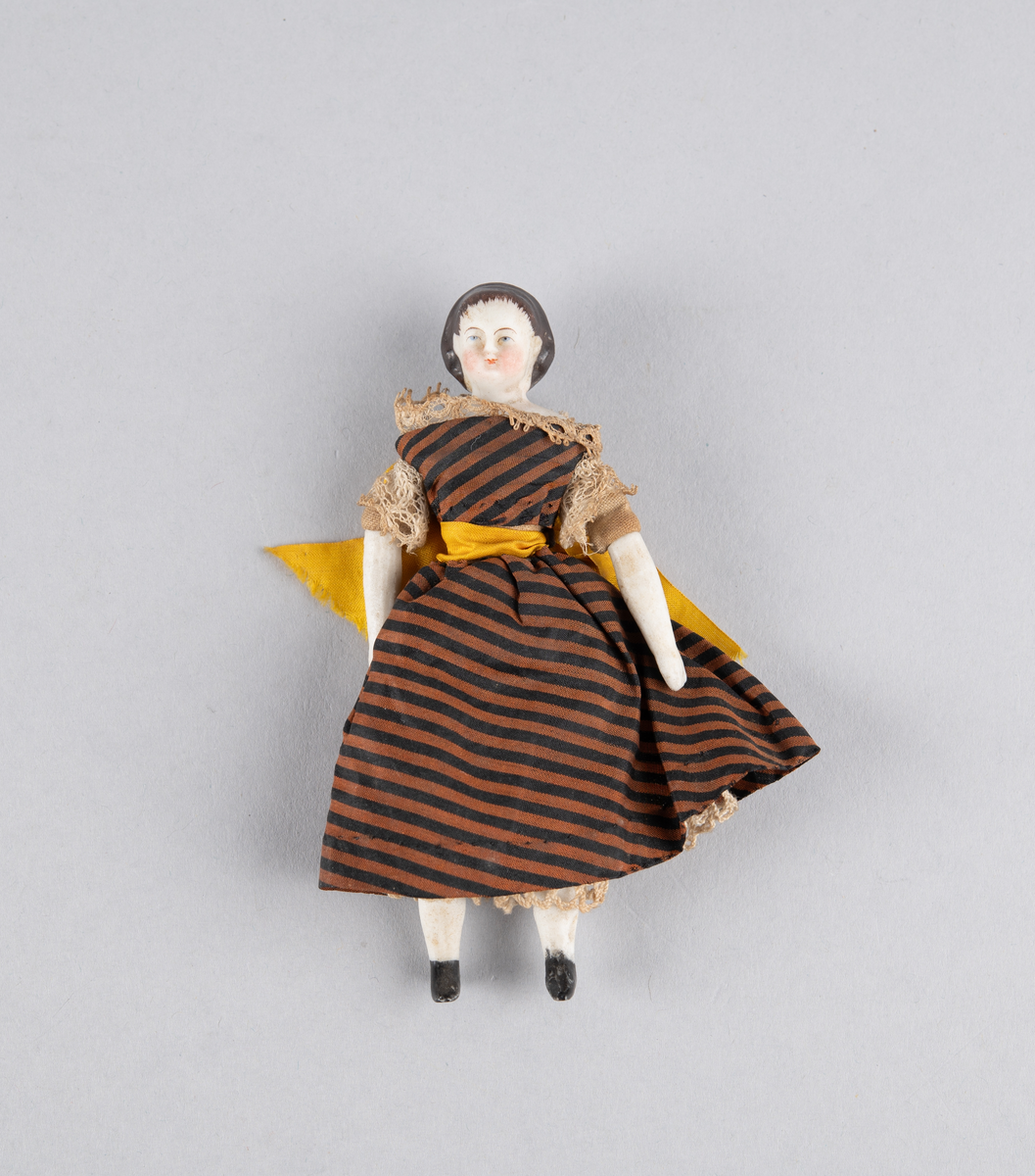 Liten porselensdukke. Kvinne med underbenklær, underskjørt og stripete silkekjole med blondekanter og sløyfe bak. Påmalte ansiktsdetaljer og støvler.