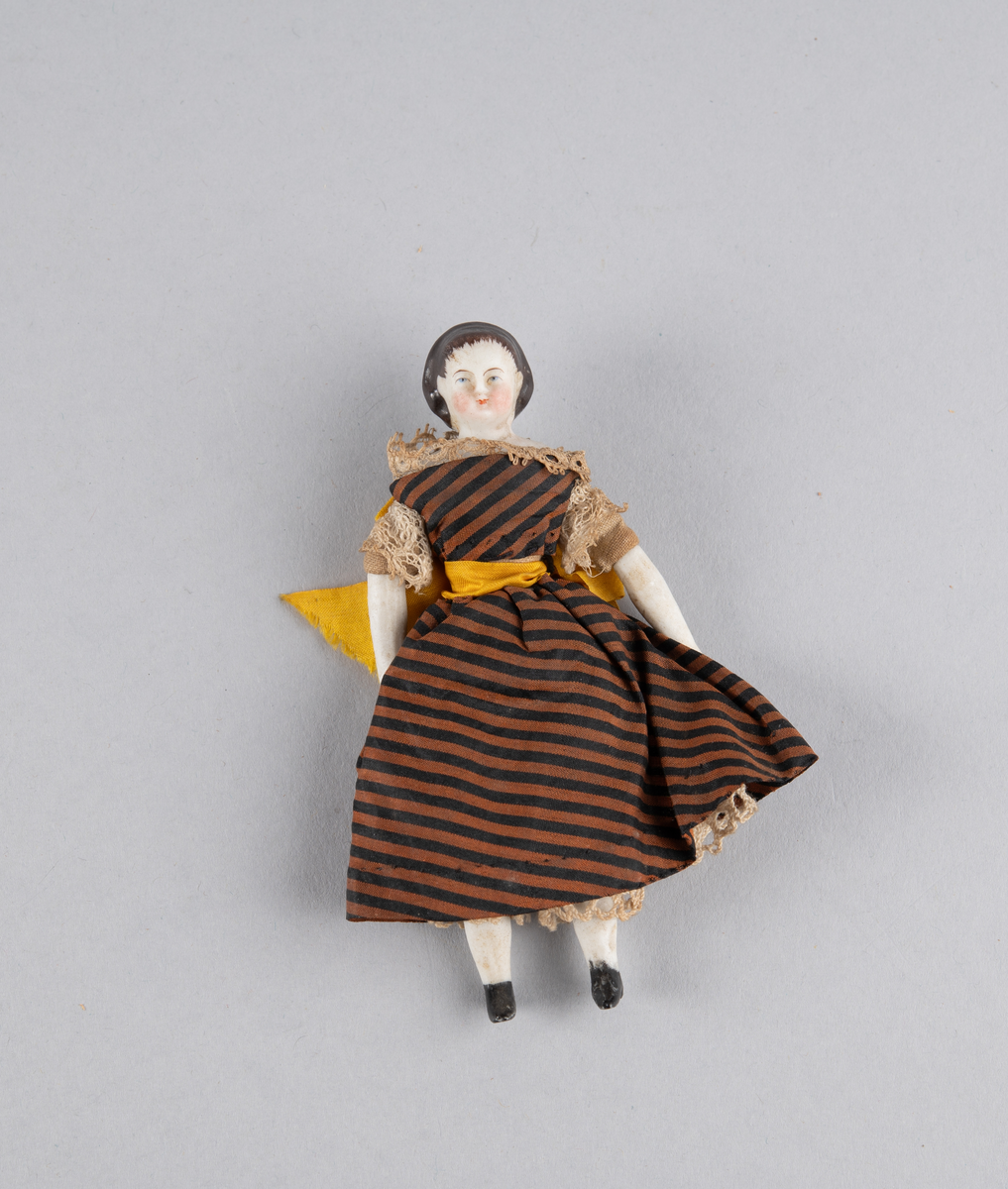 Liten porselensdukke. Kvinne med underbenklær, underskjørt og stripete silkekjole med blondekanter og sløyfe bak. Påmalte ansiktsdetaljer og støvler.
