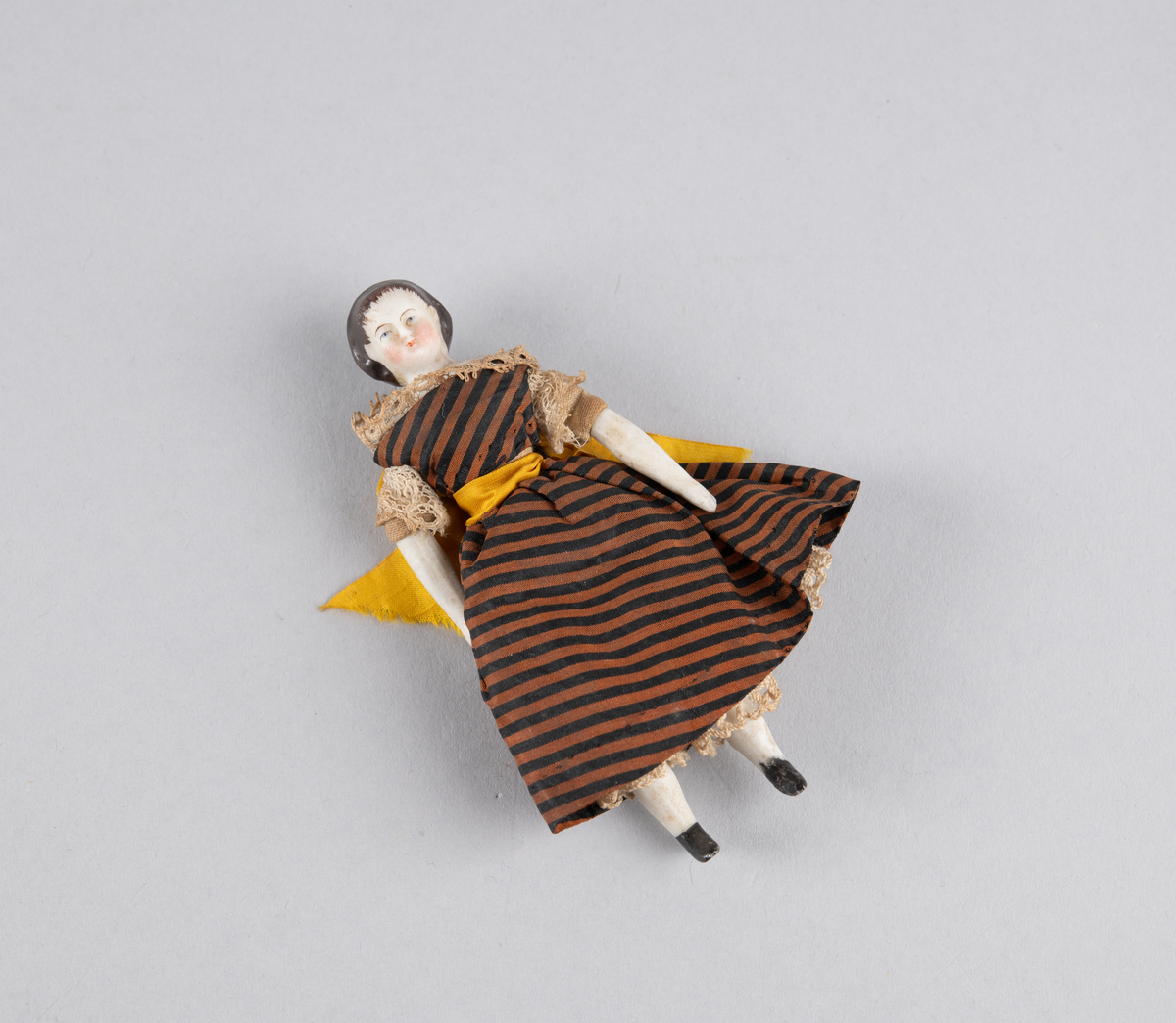 Liten porselensdukke. Kvinne med underbenklær, underskjørt og stripete silkekjole med blondekanter og sløyfe bak. Påmalte ansiktsdetaljer og støvler.