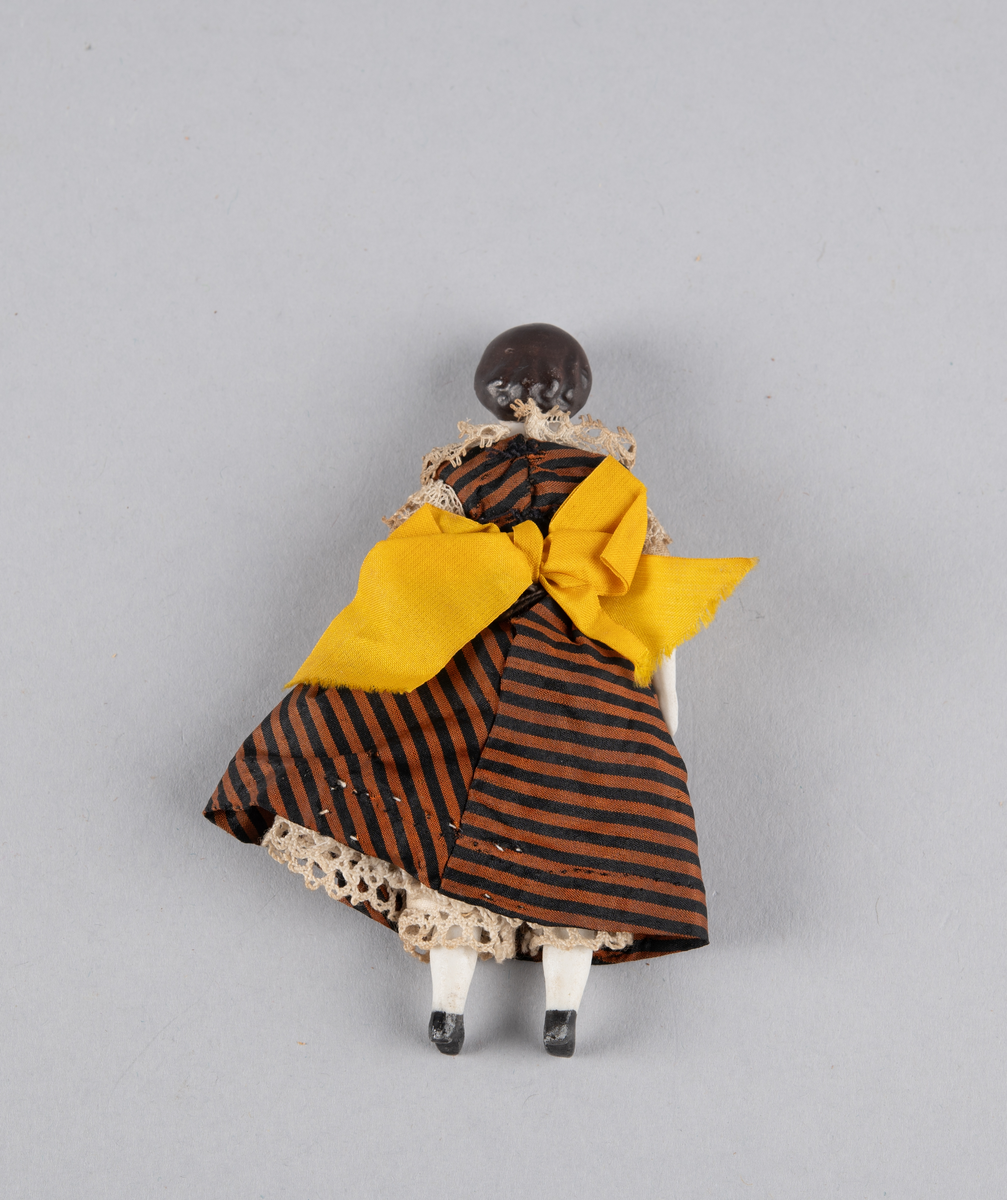 Liten porselensdukke. Kvinne med underbenklær, underskjørt og stripete silkekjole med blondekanter og sløyfe bak. Påmalte ansiktsdetaljer og støvler.