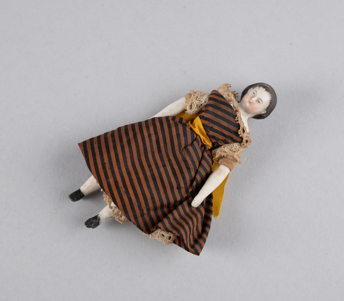 Liten porselensdukke. Kvinne med underbenklær, underskjørt og stripete silkekjole med blondekanter og sløyfe bak. Påmalte ansiktsdetaljer og støvler.