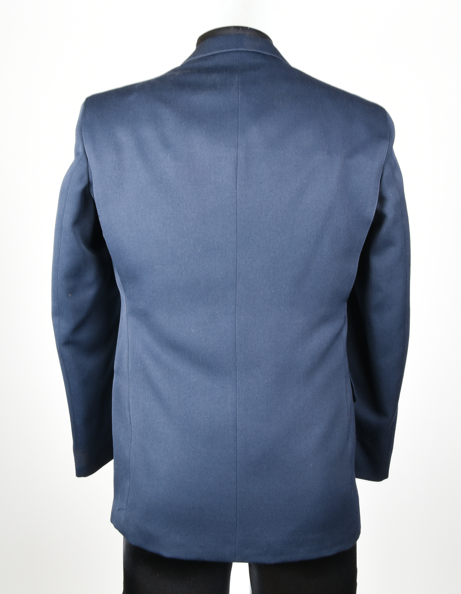 Kavaj i mörkblått tyg av ylle och polyester. Enkelknäppt med tre knappar i gulmetall. Framtill en bröstficka på vänster sida och två midjefickor, varav den på höger sida har lock. Broderat tjänstetecken vid bröstfickan. Vit lapp med svart text vid vänster ärmslut: "HERRKAVAJ VINTER C46
2110-C46
[streckkod] 930327
Ut.h. 97-02-06".
Fyra innerfickor, två på varje sida. Fastsydd vid lapp med svart text på vänster insida, mellan innerfickorna: 
"YTTERTYG
ULL, WOOL  45%
POLYESTER  55%
FODER, LINING
POLYESTER  100%
[symboler för tvättråd] Maskintvätt 40°, Varmt strykjärn, KLorblekmedel får ej användas, Endast kemtvätt med perkloreten, Får ej torktumlas.
C 46 
Johan Skoglund".
I nacken vit hank för upphängning av kavajen med svart text: "Johan Skoglund".