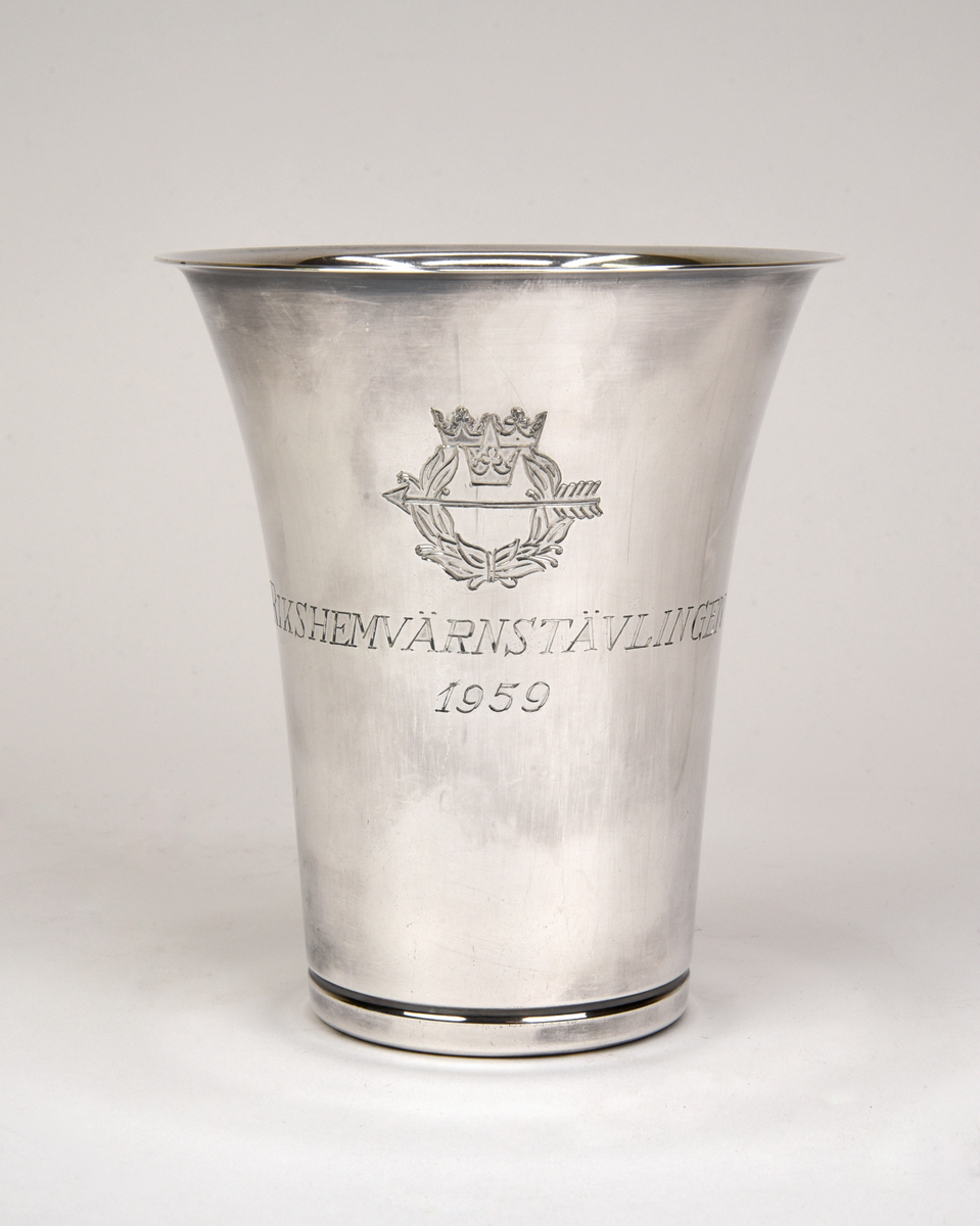 Bägare av silver. Konisk form med utvikt kant upptill. Graverad emblem och text framtill "RIKSHEMVÄRNSTÄVLINGEN 1959" och baktill "SKÄNKT AV SKYTTEFÖRBUNDENS ÖVERSTYRELSE".