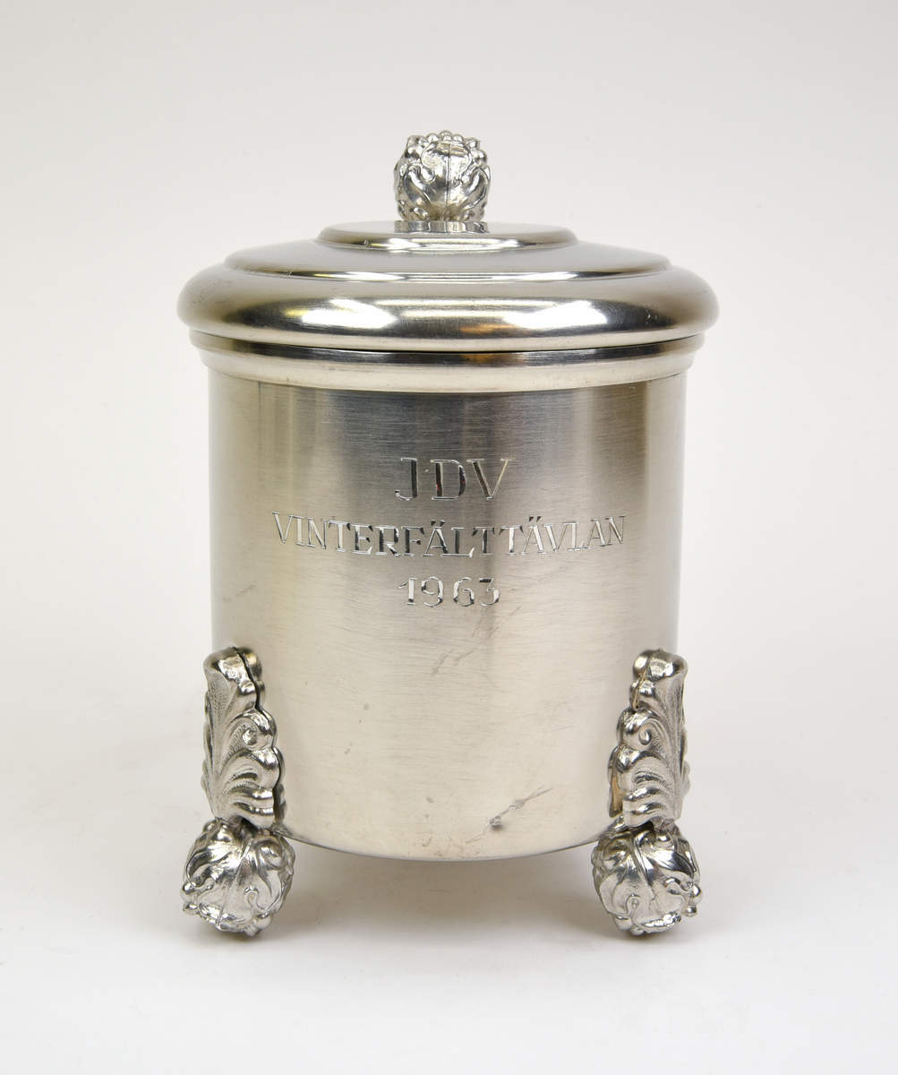 Stop av tenn med ett gängat lock. Stående på tre fötter i form av blomknoppar, en blomknopp upptill för öppning av stopet. Ornamenterad och svängd hänkel. Graverad text framtill "JDV VINTERFÄLTTÄVLAN 1963". Stämpel undertill "SVENSKT TENN C9".
