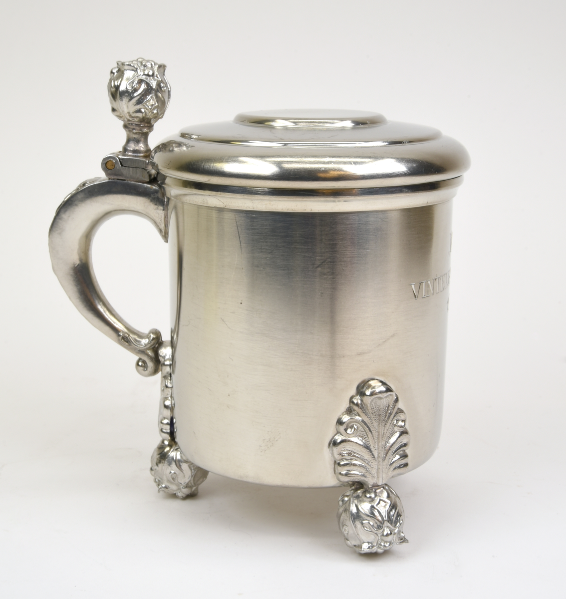 Stop av tenn med ett gängat lock. Stående på tre fötter i form av blomknoppar, en blomknopp upptill för öppning av stopet. Ornamenterad och svängd hänkel. Graverad text framtill "JDV VINTERFÄLTTÄVLAN 1963". Stämpel undertill "SVENSKT TENN C9".