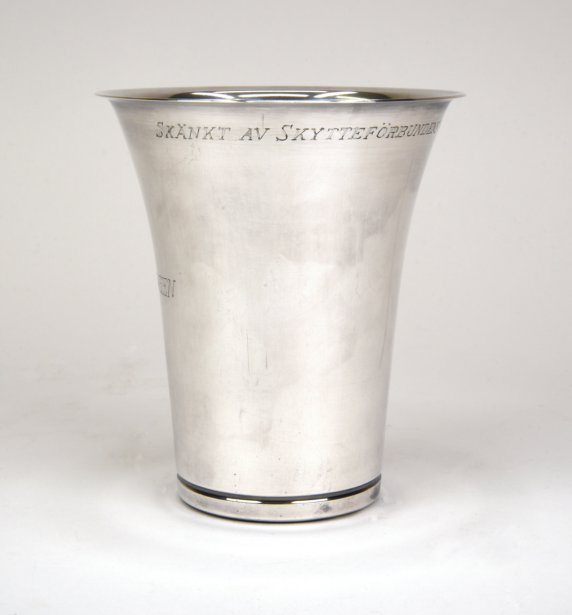 Bägare av silver. Konisk form med utvikt kant upptill. Graverad emblem och text framtill "RIKSHEMVÄRNSTÄVLINGEN 1959" och baktill "SKÄNKT AV SKYTTEFÖRBUNDENS ÖVERSTYRELSE".