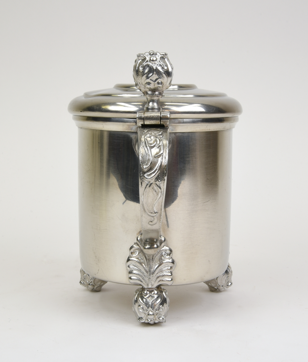 Stop av tenn med ett gängat lock. Stående på tre fötter i form av blomknoppar, en blomknopp upptill för öppning av stopet. Ornamenterad och svängd hänkel. Graverad text framtill "JDV VINTERFÄLTTÄVLAN 1963". Stämpel undertill "SVENSKT TENN C9".