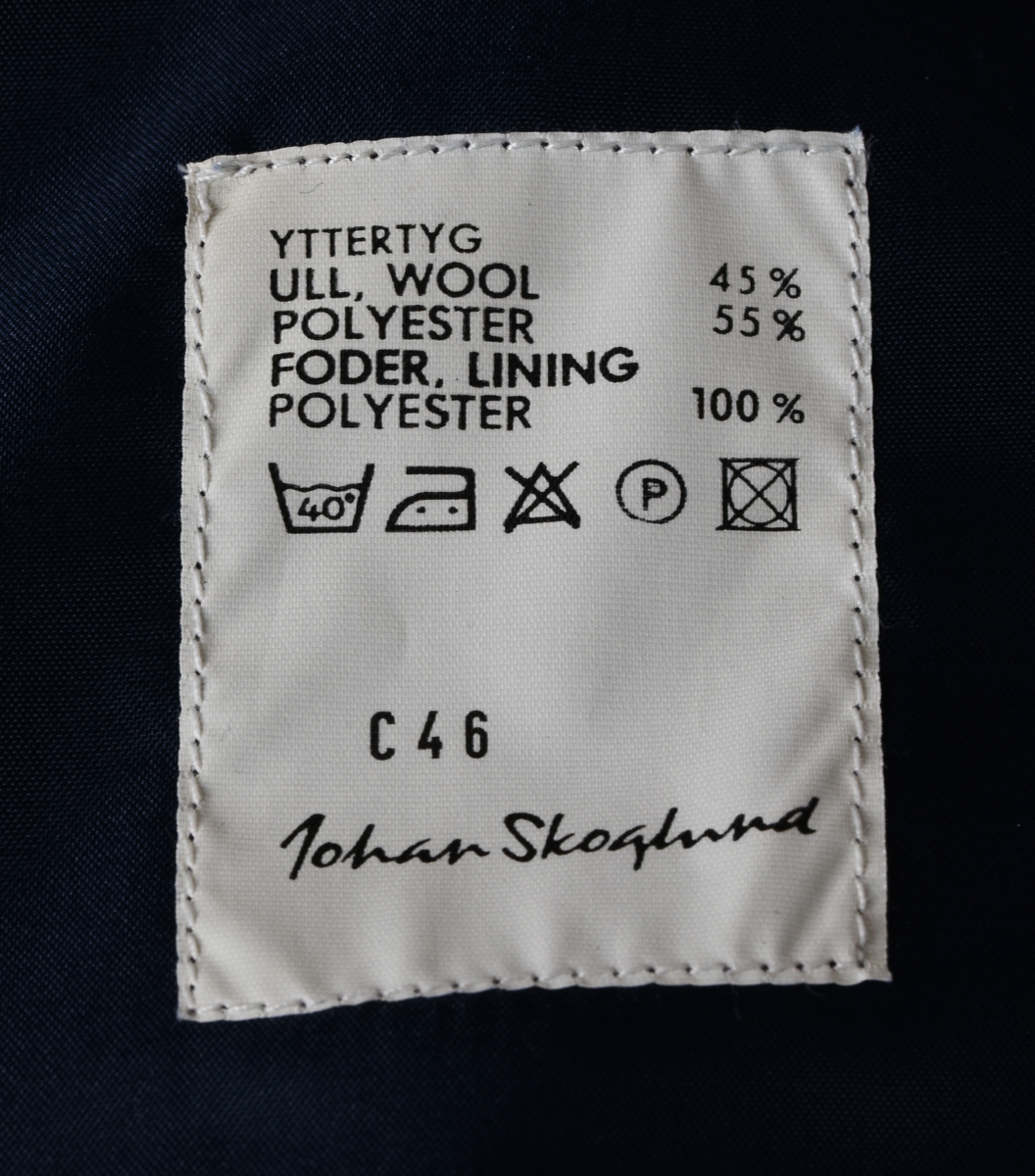 Kavaj i mörkblått tyg av ylle och polyester. Enkelknäppt med tre knappar i gulmetall. Framtill en bröstficka på vänster sida och två midjefickor, varav den på höger sida har lock. Broderat tjänstetecken vid bröstfickan. Vit lapp med svart text vid vänster ärmslut: "HERRKAVAJ VINTER C46
2110-C46
[streckkod] 930327
Ut.h. 97-02-06".
Fyra innerfickor, två på varje sida. Fastsydd vid lapp med svart text på vänster insida, mellan innerfickorna: 
"YTTERTYG
ULL, WOOL  45%
POLYESTER  55%
FODER, LINING
POLYESTER  100%
[symboler för tvättråd] Maskintvätt 40°, Varmt strykjärn, KLorblekmedel får ej användas, Endast kemtvätt med perkloreten, Får ej torktumlas.
C 46 
Johan Skoglund".
I nacken vit hank för upphängning av kavajen med svart text: "Johan Skoglund".