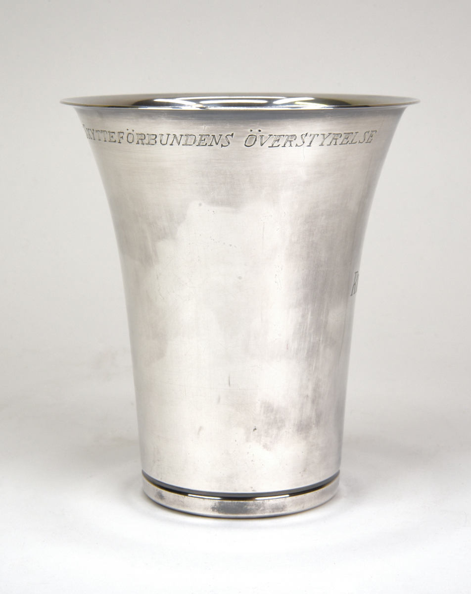 Bägare av silver. Konisk form med utvikt kant upptill. Graverad emblem och text framtill "RIKSHEMVÄRNSTÄVLINGEN 1959" och baktill "SKÄNKT AV SKYTTEFÖRBUNDENS ÖVERSTYRELSE".