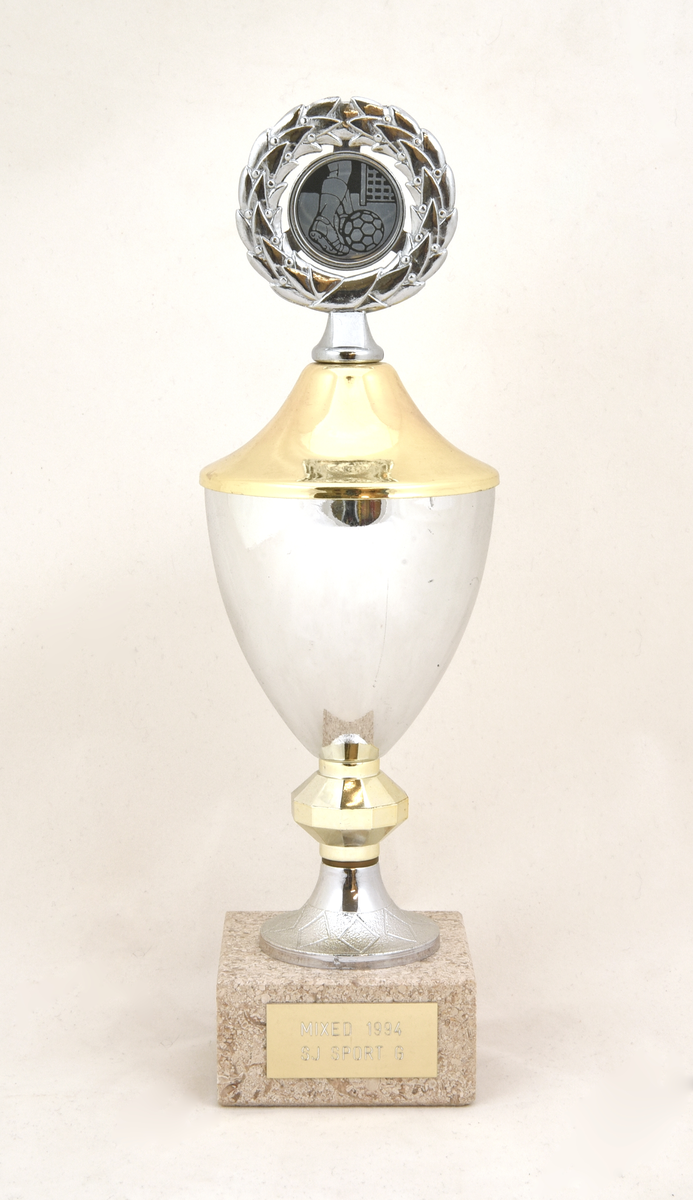 Pokal (:1) med guldfärgat lock (:2), och fot av grå stengods. På locket upptill en lagerkrans med en illustration i form av en fotbollsspelares ben som skjuter bollen mot mål. Pokalens överdel består av en silverfärgad metallskål som är guldfärgad på insidan. Fastlimmad guldfärgad metallbricka på foten med text "MIXED 1994 SJ SPORT G". Det finns ytterligare en etikett på pokalens undersida vars ytskikt är bortnött.