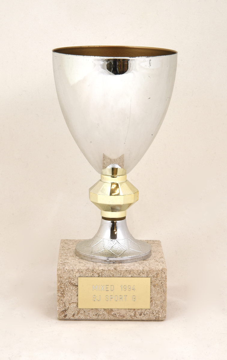 Pokal (:1) med guldfärgat lock (:2), och fot av grå stengods. På locket upptill en lagerkrans med en illustration i form av en fotbollsspelares ben som skjuter bollen mot mål. Pokalens överdel består av en silverfärgad metallskål som är guldfärgad på insidan. Fastlimmad guldfärgad metallbricka på foten med text "MIXED 1994 SJ SPORT G". Det finns ytterligare en etikett på pokalens undersida vars ytskikt är bortnött.