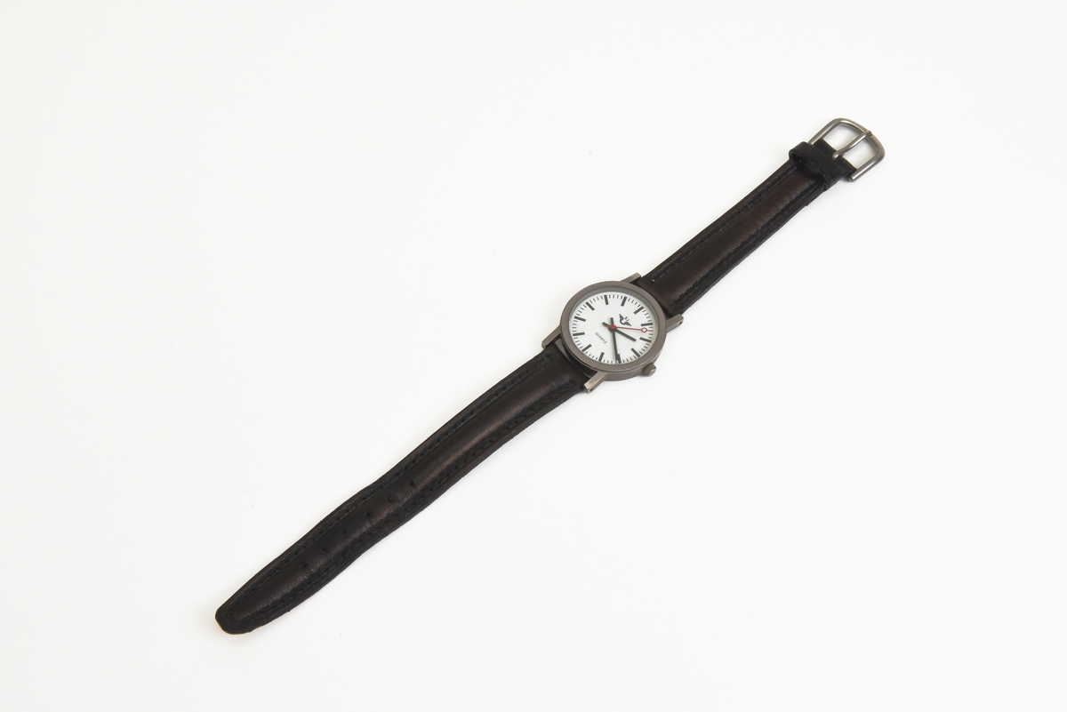 Armbandsur (:1) i blå förvaringsask (:2).

Runt armbandsurets inglasade urtavla löper en grå metallram. Bandet är tillverkat av svart skinn. Urtavlan är vit med svarta detaljer samt en röd sekundvisare. På urtavlan syns texten QUARTZ samt SJ:s firmamärke i svart. Längst ut på bandet sitter ett spänne i grå metall.
Förvaringsasken är av blå plast. På utsidan finns SJ:s firmamärke i "guldfärg". Askens insida är klädd med grå textil.