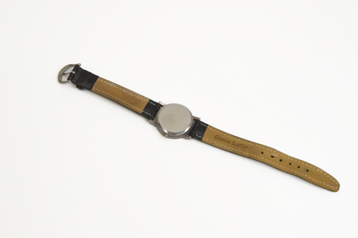 Armbandsur (:1) i blå förvaringsask (:2).

Runt armbandsurets inglasade urtavla löper en grå metallram. Bandet är tillverkat av svart skinn. Urtavlan är vit med svarta detaljer samt en röd sekundvisare. På urtavlan syns texten QUARTZ samt SJ:s firmamärke i svart. Längst ut på bandet sitter ett spänne i grå metall.
Förvaringsasken är av blå plast. På utsidan finns SJ:s firmamärke i "guldfärg". Askens insida är klädd med grå textil.