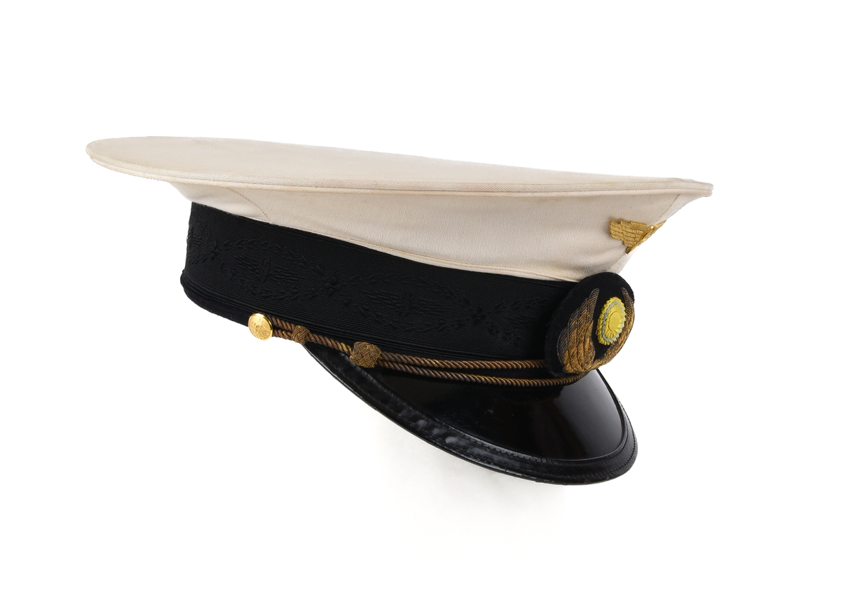 Skärmmössa av 1921 års reglemente, för högre tjänsteman vid enskild järnväg. Mössan har vitt sommarkapell. Framtill på kapellet sitter ett tjänstetecken i form av ett guldfärgat vinghjul av metall. Bandet är svart och med svart dekor föreställande vinghjul och växtornamentik. Tränsen utgörs av en guldfärgad snodd fäst i två guldfärgade knappar vid tinningen. Knapparna har motiv föreställande vinghjul med åskviggar. Framtill på bandet sitter ett textilt tjänstetecken med vinghjul och nationalkokard. Mössan har svart lackskärm. 
Skärmen är grön på insidan. Fodret är vitt och svettbandet gråblått. I fodret återfinns namnet på tillverkaren, Nymarks Norrköping.