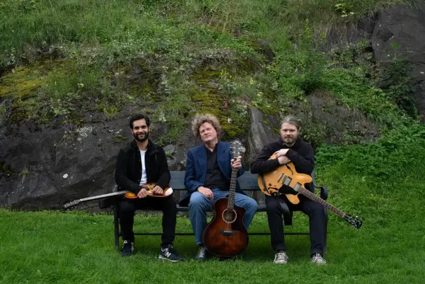 Trioen Haakon Ellingsen, Jacob Holthe Thorsen og Gaute Storsve sitter på benk med gitarer foran grønn skråning.