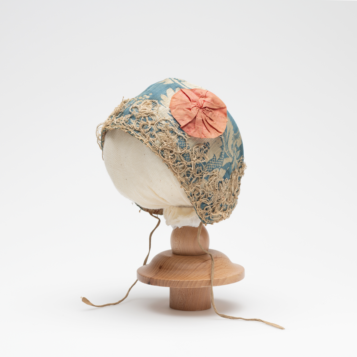 Dåpslue til gutt av slike, hvit mønster på blå bunn. Seks deler sydd sammen. Dekoret langs kanten med blonde i uidentifisert teknikk. Dekorert med to rød og rosa blomster, den røde sjuler punktet der de seks sømmen møtes. Blomstene er antagelig sekundære. Innvendig foret med bomullstoff, rød og blå striper på hvit bunn. Enkel hvit bomullsnor på hver side.