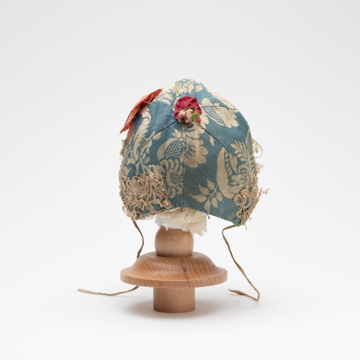 Dåpslue til gutt av slike, hvit mønster på blå bunn. Seks deler sydd sammen. Dekoret langs kanten med blonde i uidentifisert teknikk. Dekorert med to rød og rosa blomster, den røde sjuler punktet der de seks sømmen møtes. Blomstene er antagelig sekundære. Innvendig foret med bomullstoff, rød og blå striper på hvit bunn. Enkel hvit bomullsnor på hver side.