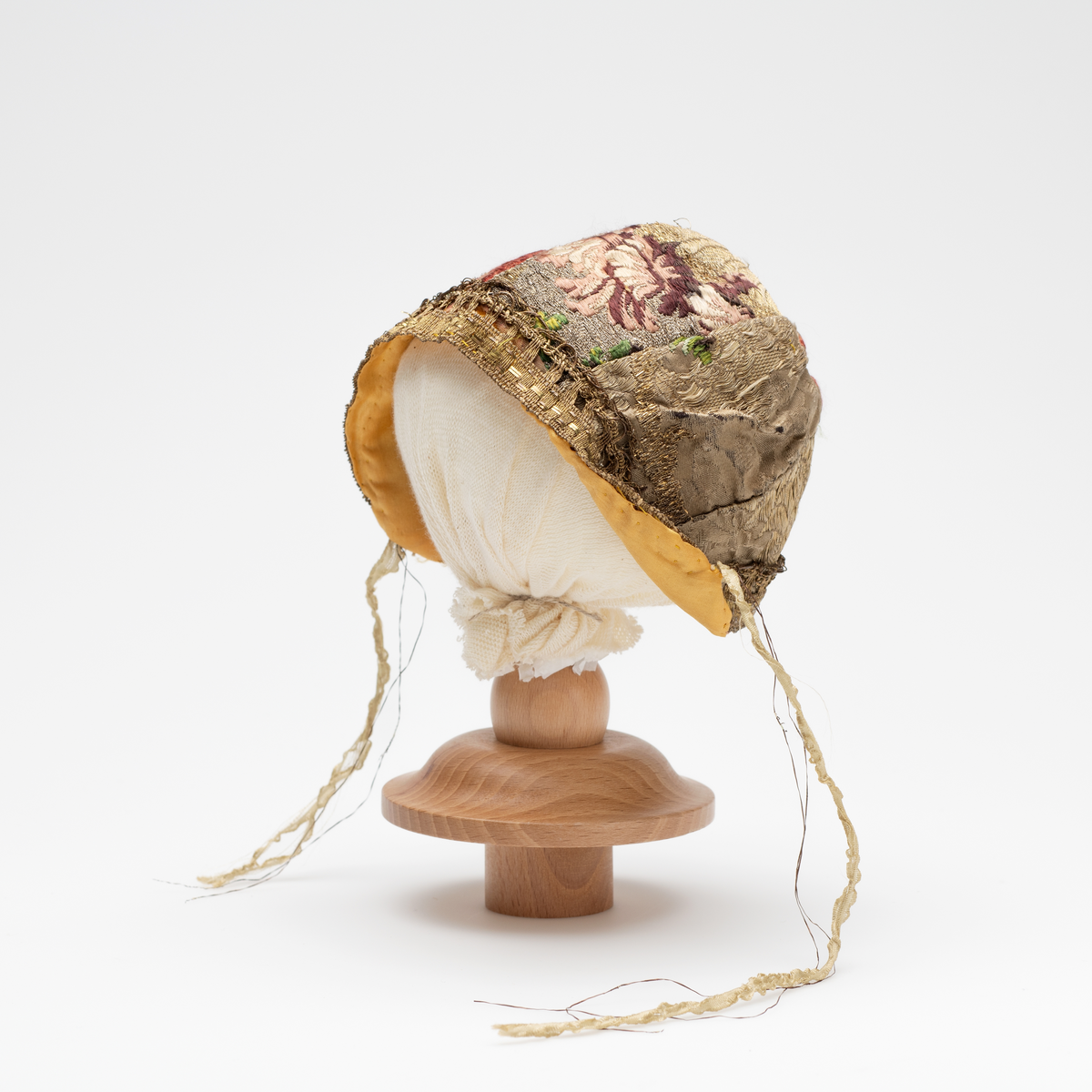 Dåpslue av slike, planterdekor i rødt, grønt og lille på sølvfarget bakgrunn. Dekorert langs kanten med gullfarget blonde i uidentifisert teknik.  Innvendig foret med gul ensfarget silke. Silkebånd på hver side. Lua er sydd sammen av åtte biter.