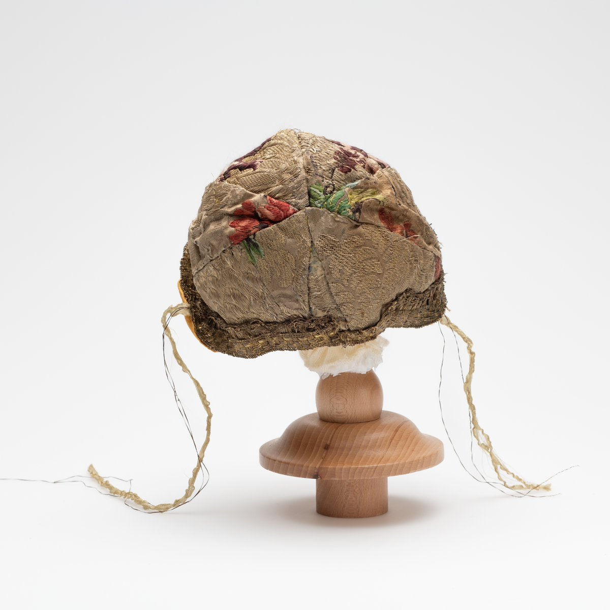 Dåpslue av slike, planterdekor i rødt, grønt og lille på sølvfarget bakgrunn. Dekorert langs kanten med gullfarget blonde i uidentifisert teknik.  Innvendig foret med gul ensfarget silke. Silkebånd på hver side. Lua er sydd sammen av åtte biter.