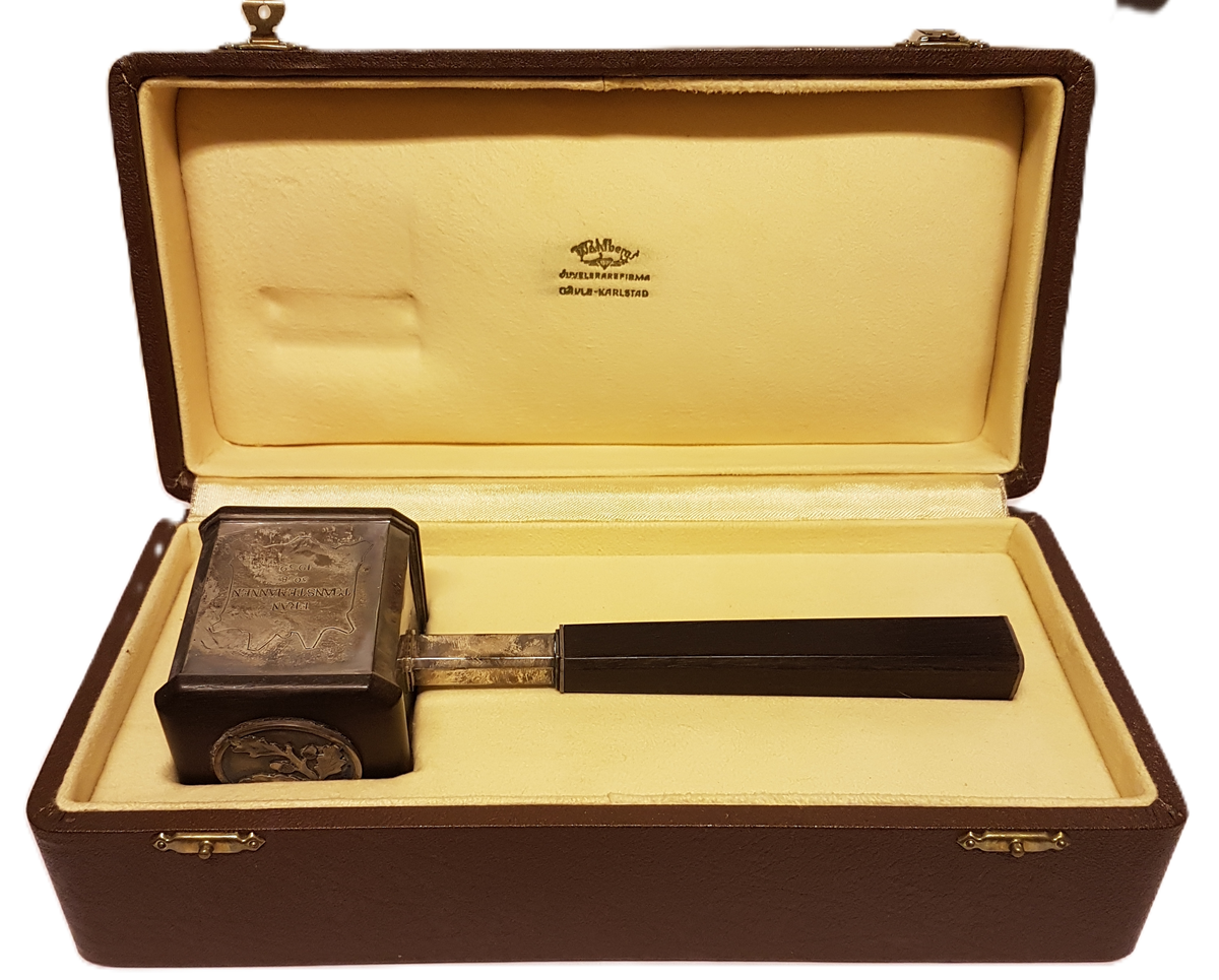 Ordförandeklubba i silver och trä. Ligger i tillhörande etui.  Inskription LA Mattons läderfabrik AB, Gefle. Från tjänstemännen 30/8 1952. Inuti locket står tryckt: Wåhlbergs juvelerarfirma Gävle-Karlstad.
