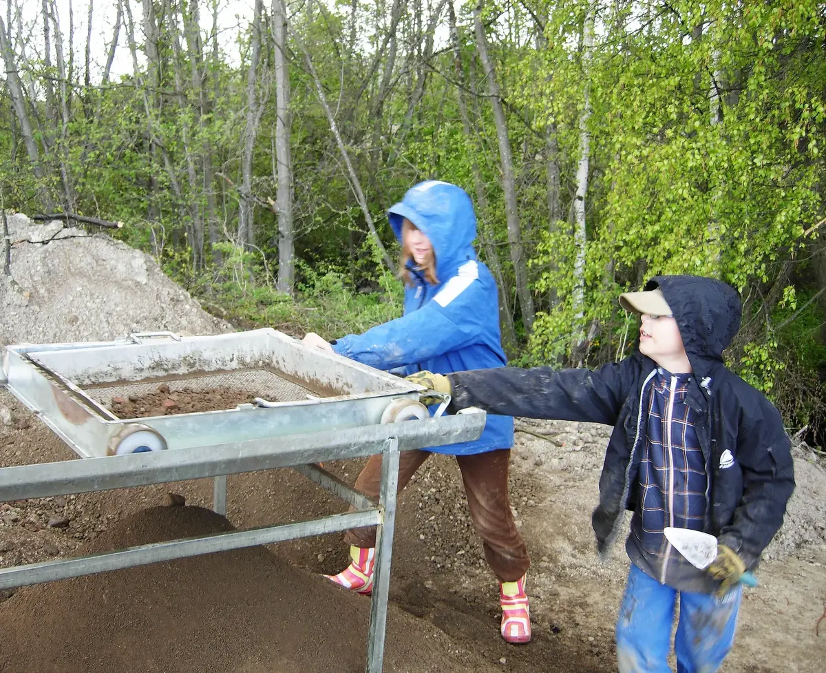 Två barn med regnkläder som står och sållar i en utgrävning med träd i bakgrunden.