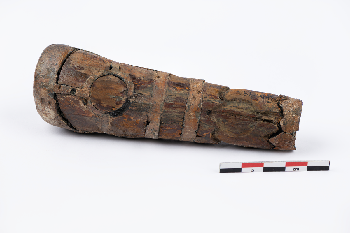 løvtre. tinn. ovalt tverrsnitt. smalner mot falen. beslått med innlagt tinn i mønster. på hver side er i øvre halpart en sirkel hvorfra 4 bånd går ut til rammen. i nedre felt en tulipan med stengel. falen har tenner på oversiden. hullet for tangen firkantet bredde 0,55 lengde 0,7.