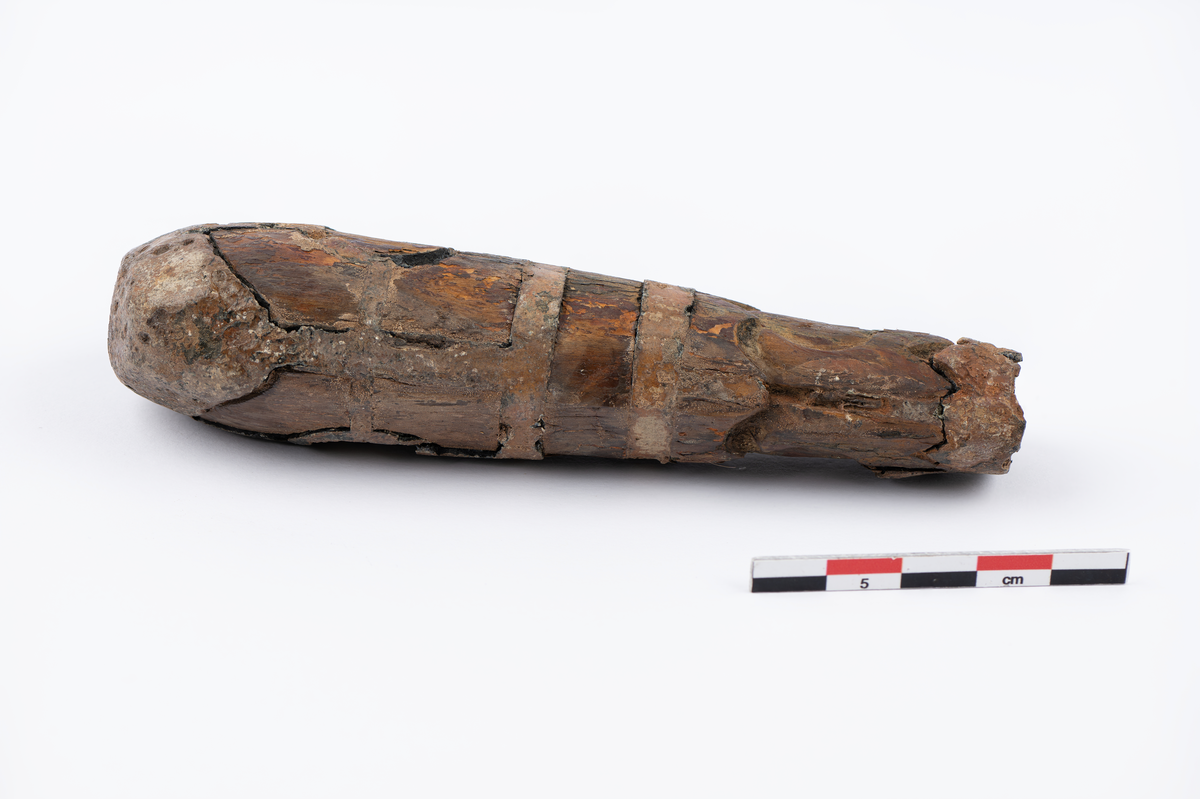 løvtre. tinn. ovalt tverrsnitt. smalner mot falen. beslått med innlagt tinn i mønster. på hver side er i øvre halpart en sirkel hvorfra 4 bånd går ut til rammen. i nedre felt en tulipan med stengel. falen har tenner på oversiden. hullet for tangen firkantet bredde 0,55 lengde 0,7.