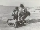 Tre man ur FBU-kurs lyfter sårad person med bandage på sandstrand, 1957.