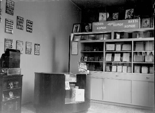 Interiör från Märta Ågrens fotobutik Fototjänst på Borgmästaregatan, där hon även hade fotoateljé. Hon startade 1935 på gatuadress nr 12, men flyttade 1940 till nr 14. Här ses montrar, hyllor och diskar med kameror, ramar och andra tillbehör. På väggarna hänger exempel på s k storbilder, eller Polyfoto, dvs många små porträtt på samma ark. (Se även bildnr B 1087_1-2)