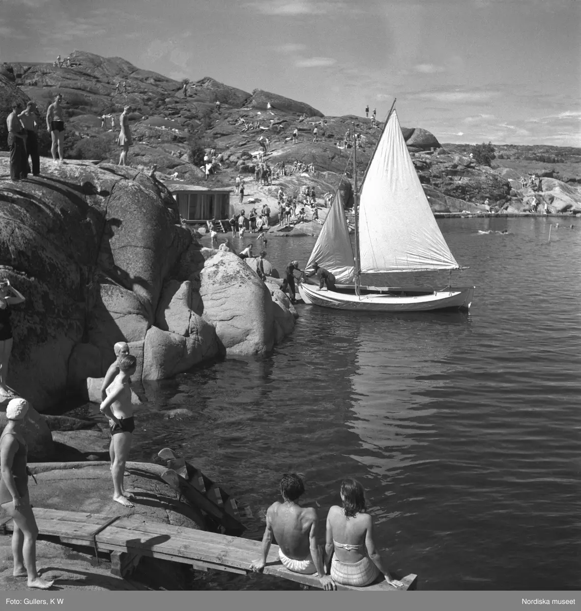 Sommarmotiv, segelbåt, badande människor, brygga, klippor och hav på västkusten.