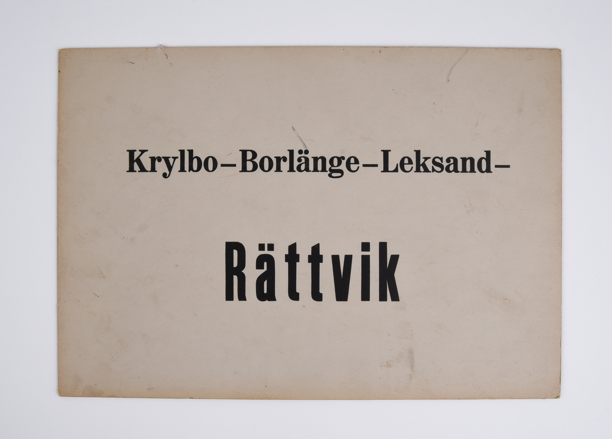 Destinationsskylt av kartong med text i svart "Krylbo - Borlänge - Leksand - Rättvik" på ena sidan och "Rättvik - Leksand - Borlänge - Krylbo" på andra sidan.