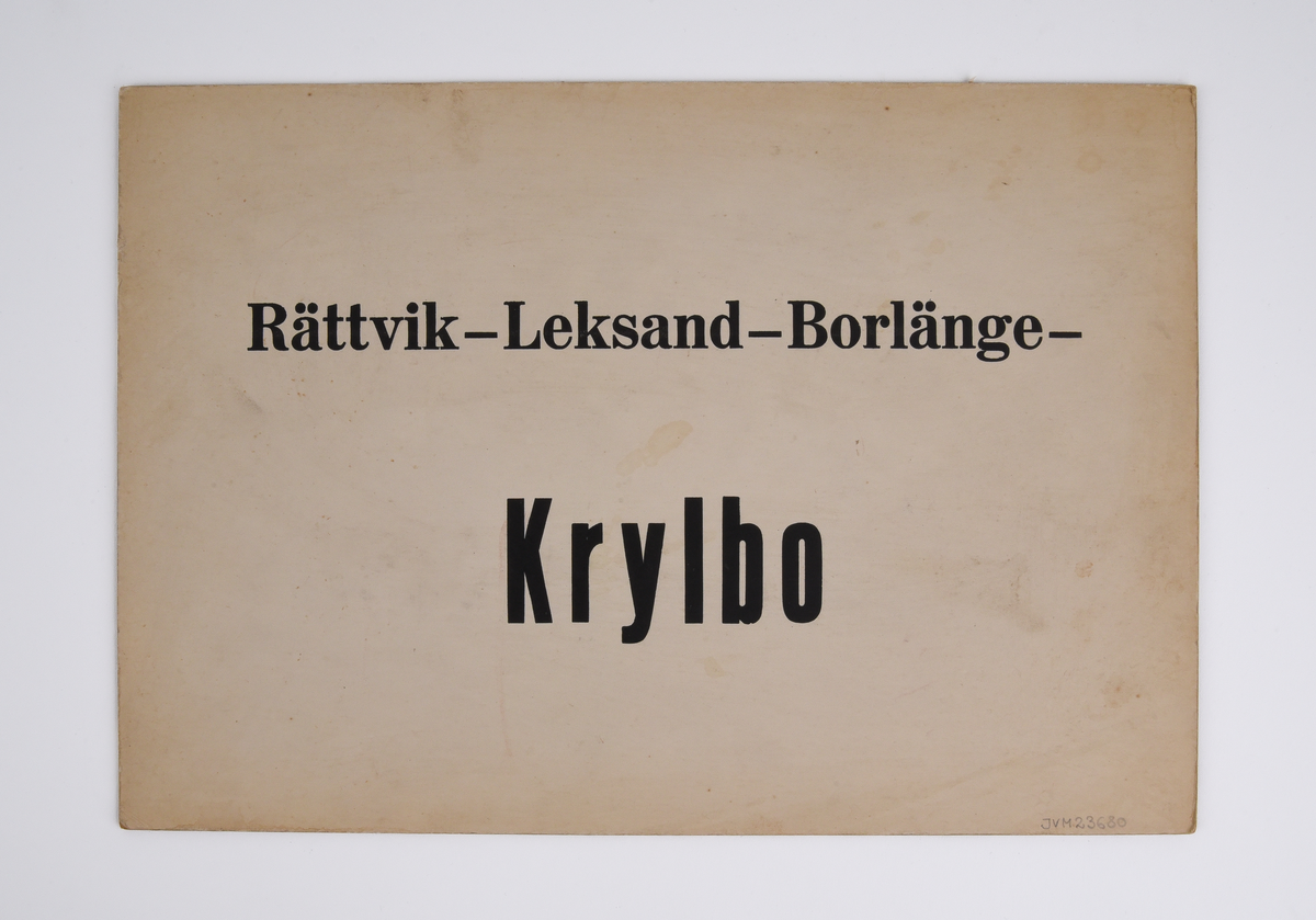 Destinationsskylt av kartong med text i svart "Krylbo - Borlänge - Leksand - Rättvik" på ena sidan och "Rättvik - Leksand - Borlänge - Krylbo" på andra sidan.