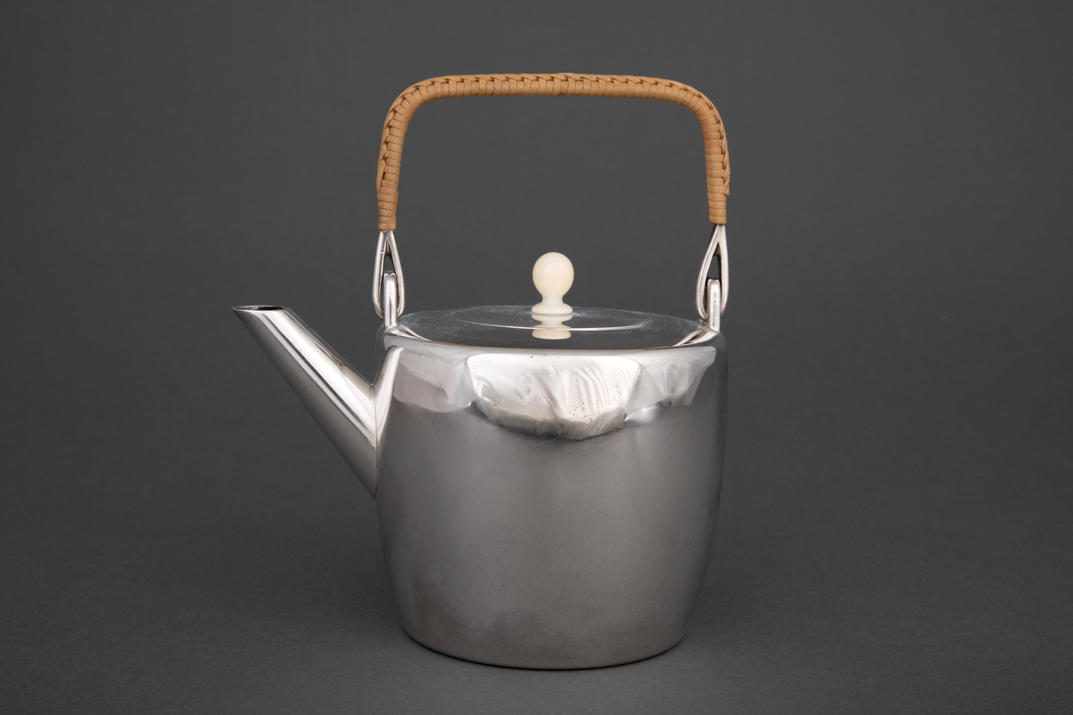 Tekanne i sølv. Sylinderformet korpus, hvorfra det reiser seg et rettvinklet håndtak med kurvfletting. På toppen er det et sirkulært lokk med lokknapp i bein, og kannens tut løper diagonalt ut fra korpus.
