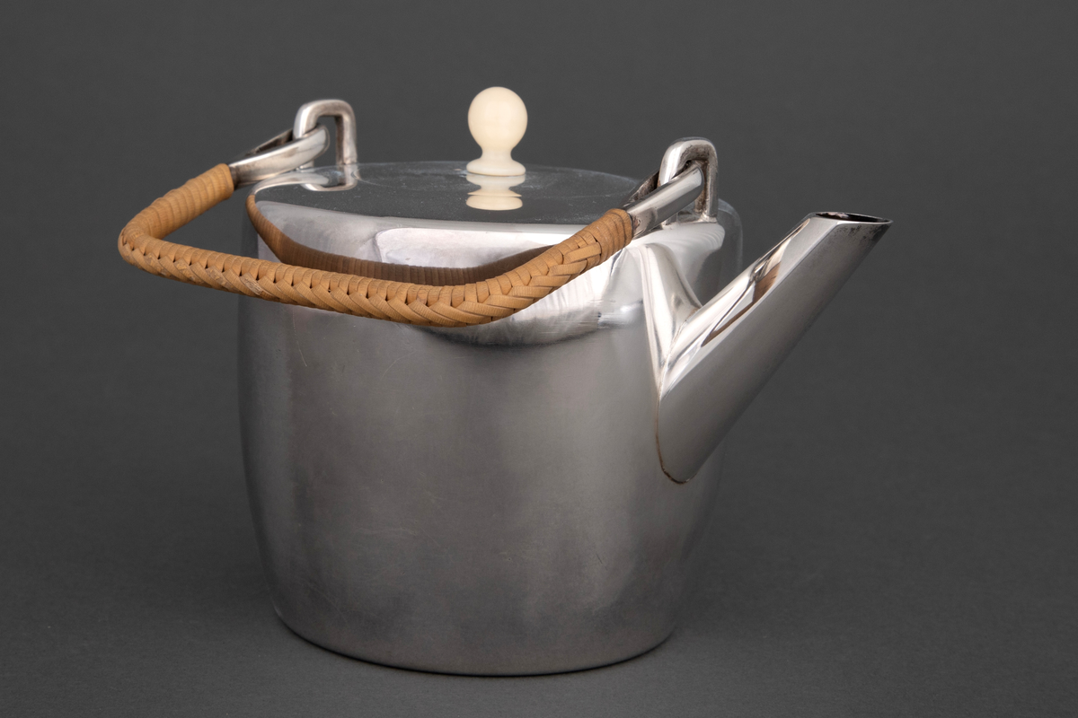 Tekanne i sølv. Sylinderformet korpus, hvorfra det reiser seg et rettvinklet håndtak med kurvfletting. På toppen er det et sirkulært lokk med lokknapp i bein, og kannens tut løper diagonalt ut fra korpus.